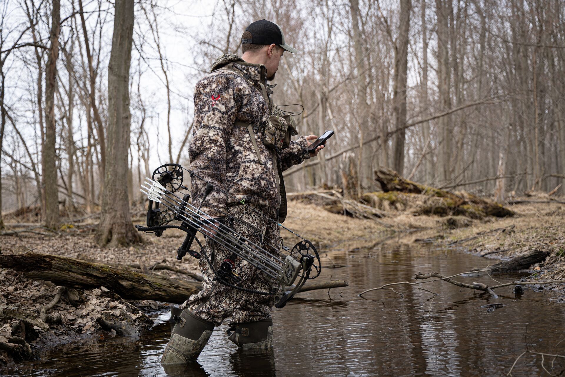 Hunter in camo walks through creek with bow and phone.