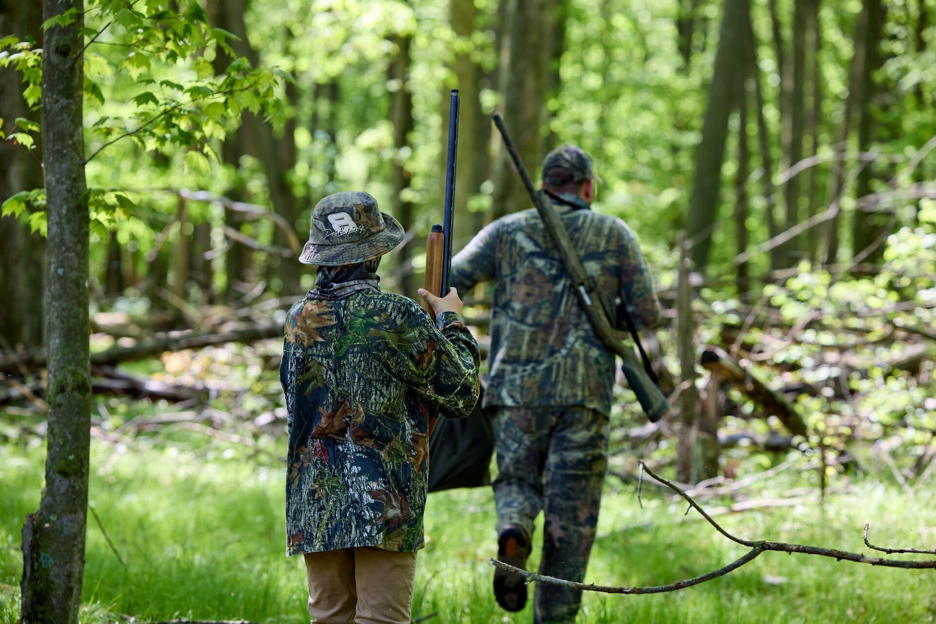 A man and boy in hunting camo with shotguns walk in woods.