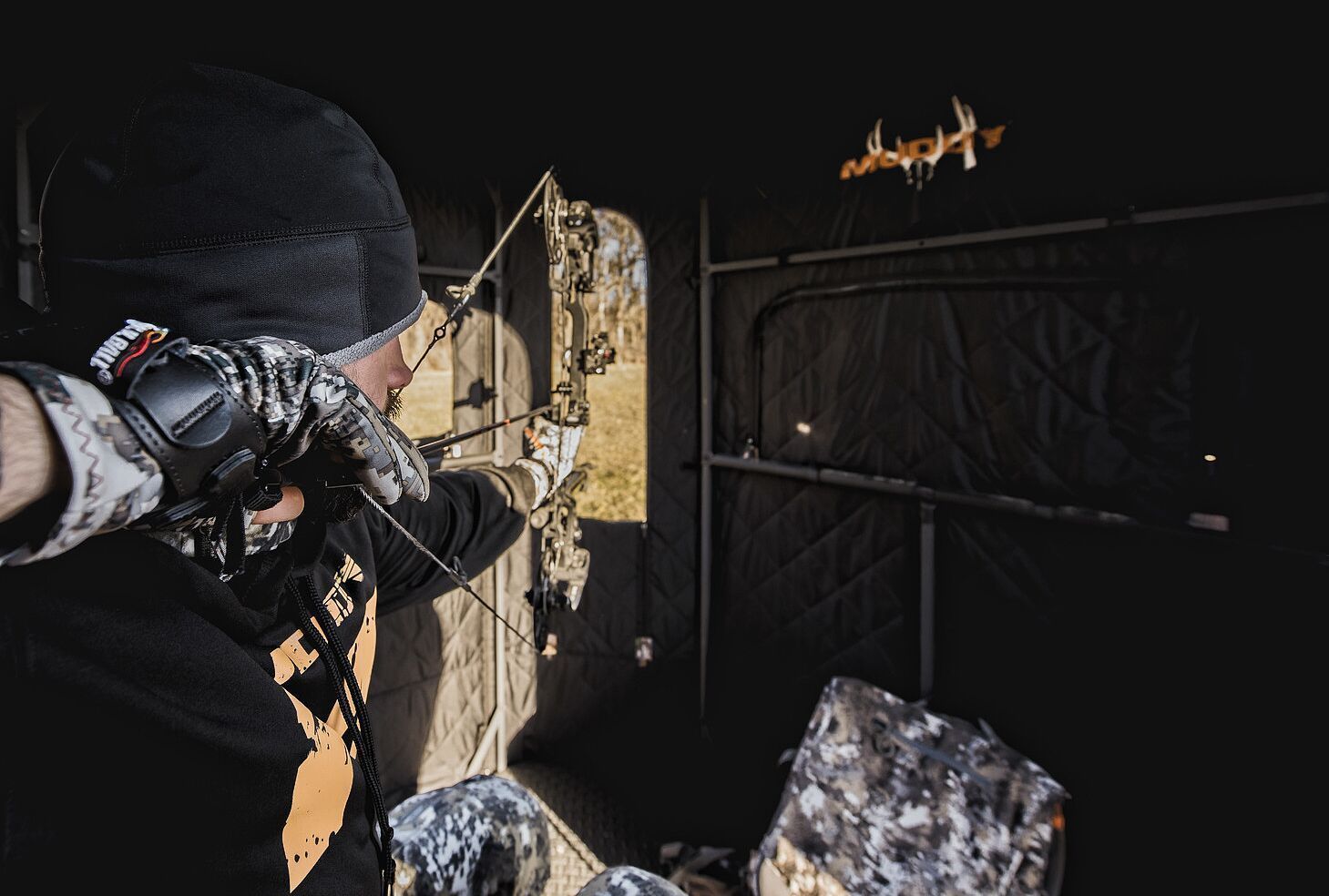 A hunter in a blind draws back his bow, choosing a bow case concept.