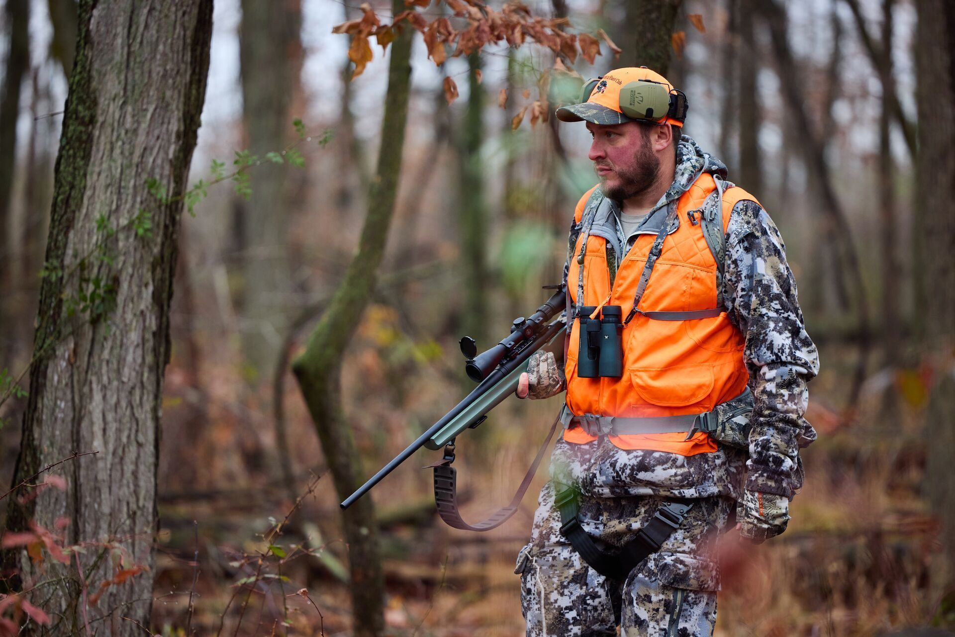 A hunter in camo and blaze orange carries a rifle in the woods.