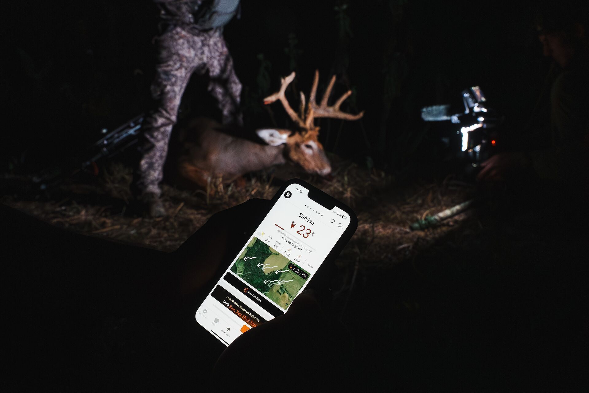 Hunter holding phone showing HuntCast with deer in background.