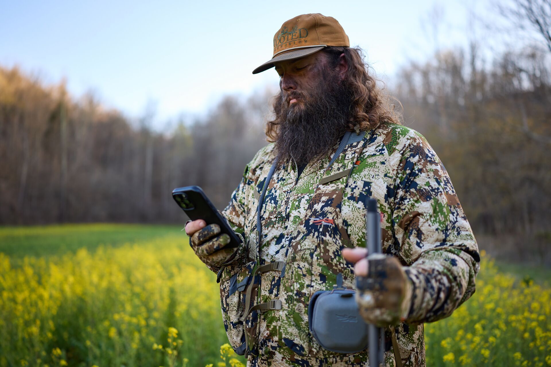 RootedTV hunter in camo with shotgun looks at phone for HuntWise hunting app.