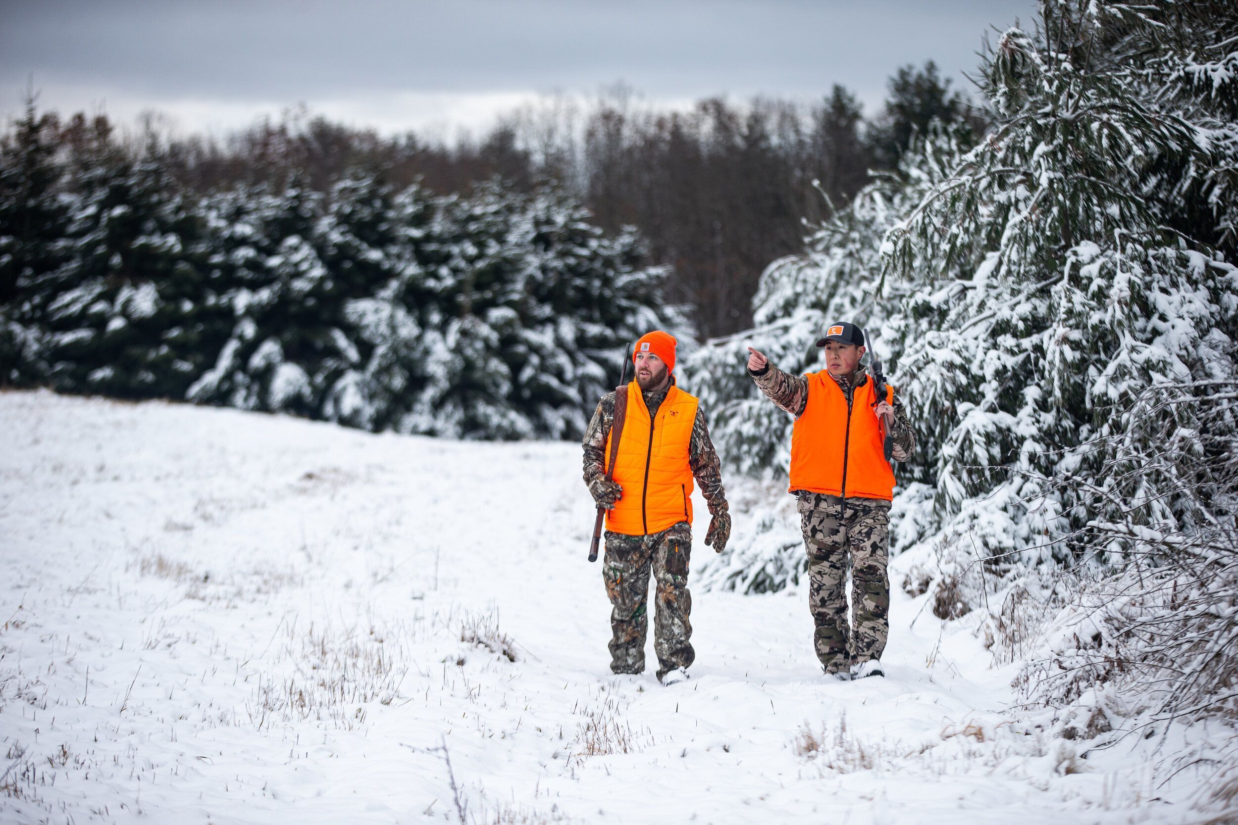Two hunters in blaze orange and camo walk through snow for late-season hunting.