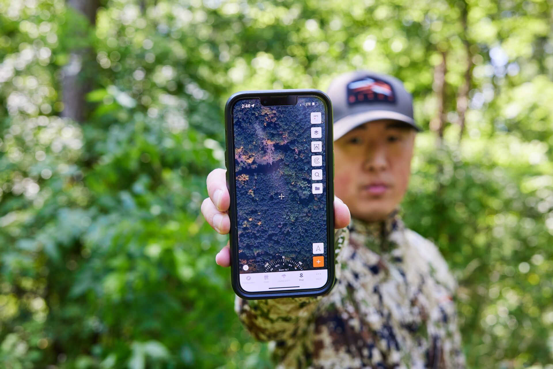 A hunter in the woods holds a phone up to the camera, showing HuntWise hunting app for deer hunts.