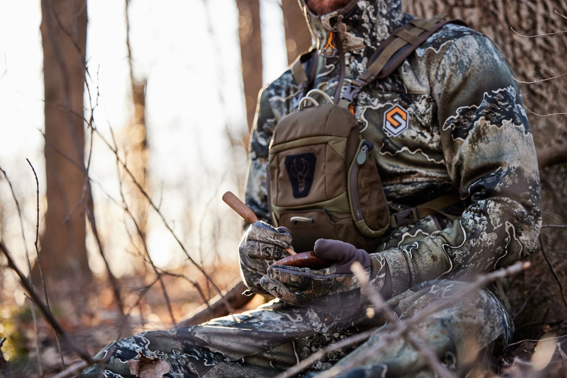 A hunter sitting against a tree in camo using a turkey call, turkey hunting PA concept.