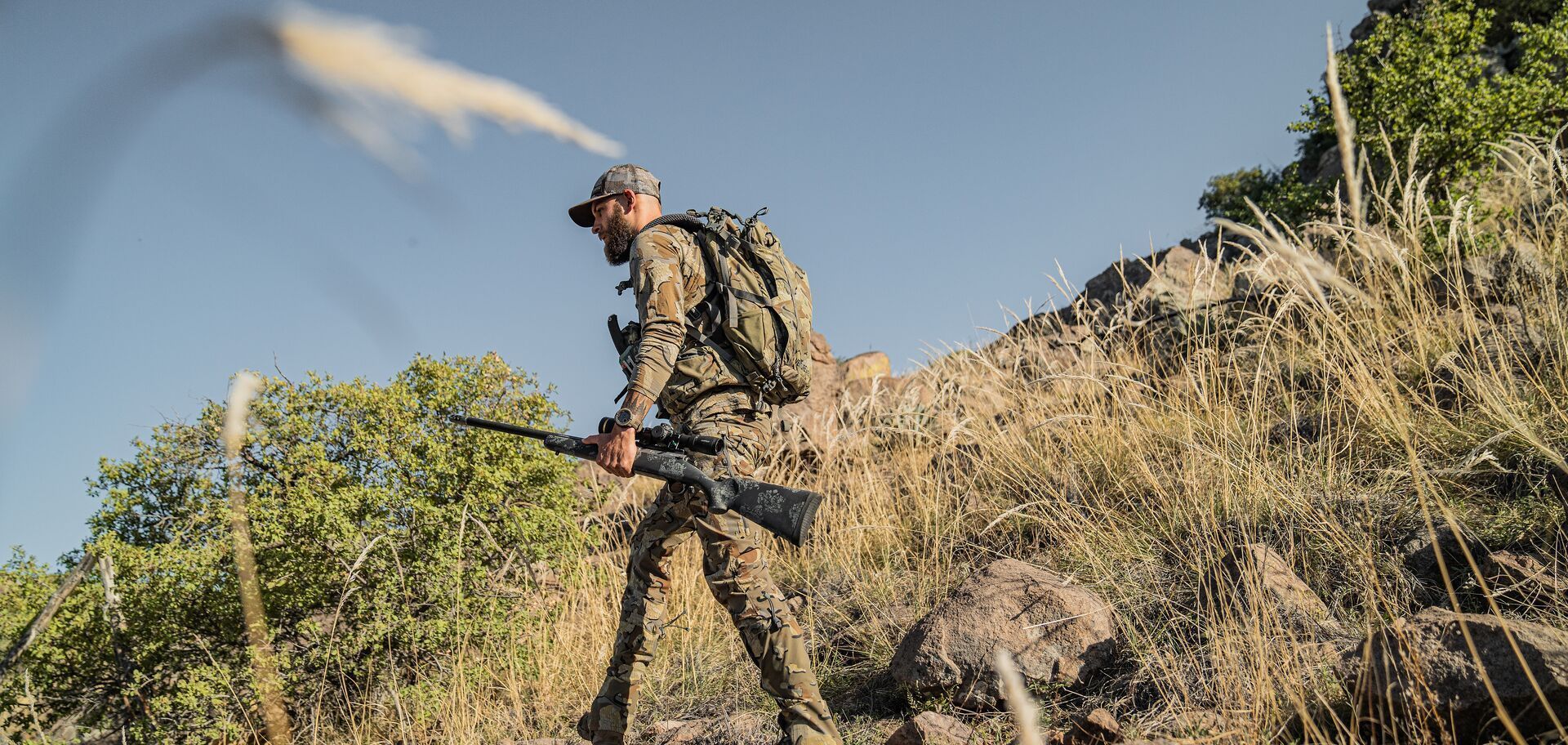Hunter in camo with rifle walks hill, mule deer season concept.