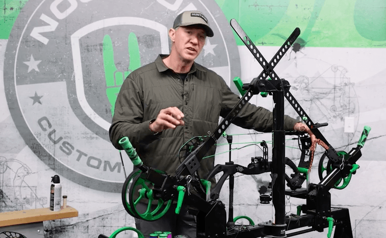 John Dudley stands behind a compound bow on a rack to talk about maintenance.