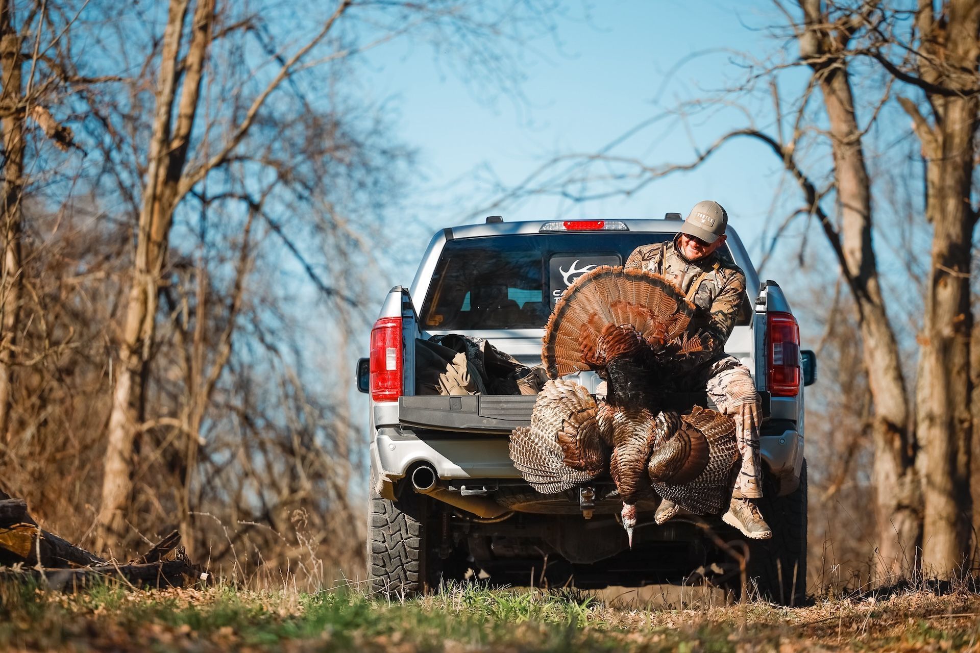 A hunter sits on the back of a truck with a wild turkey after a hunt.