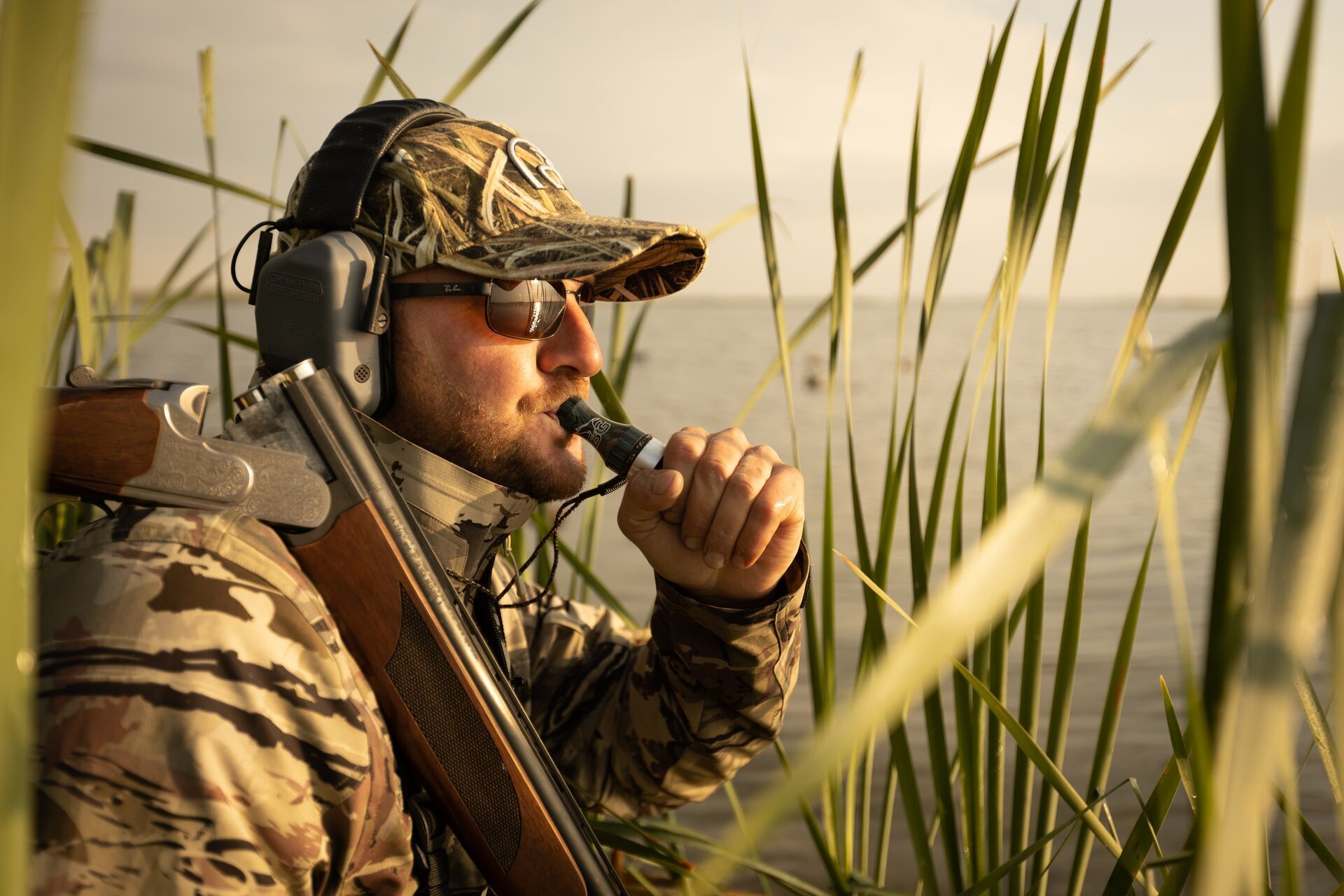 A hunter with a shotgun using a duck call.
