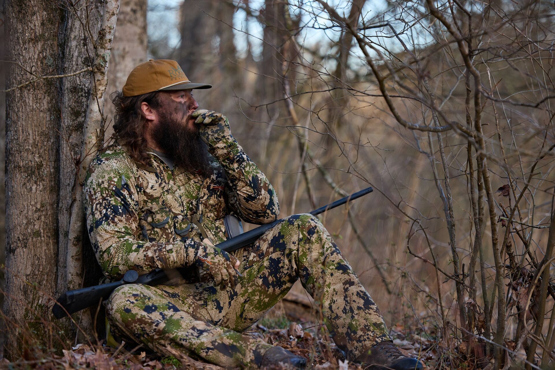 RootedTV hunters in camo leans against a tree and uses turkey call, turkey hunting tips concept.