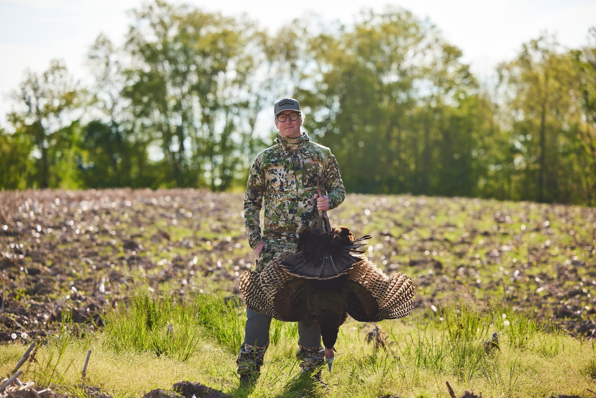 A smiling hunter in camo holds up a turkey kill, represents Indiana turkey season.