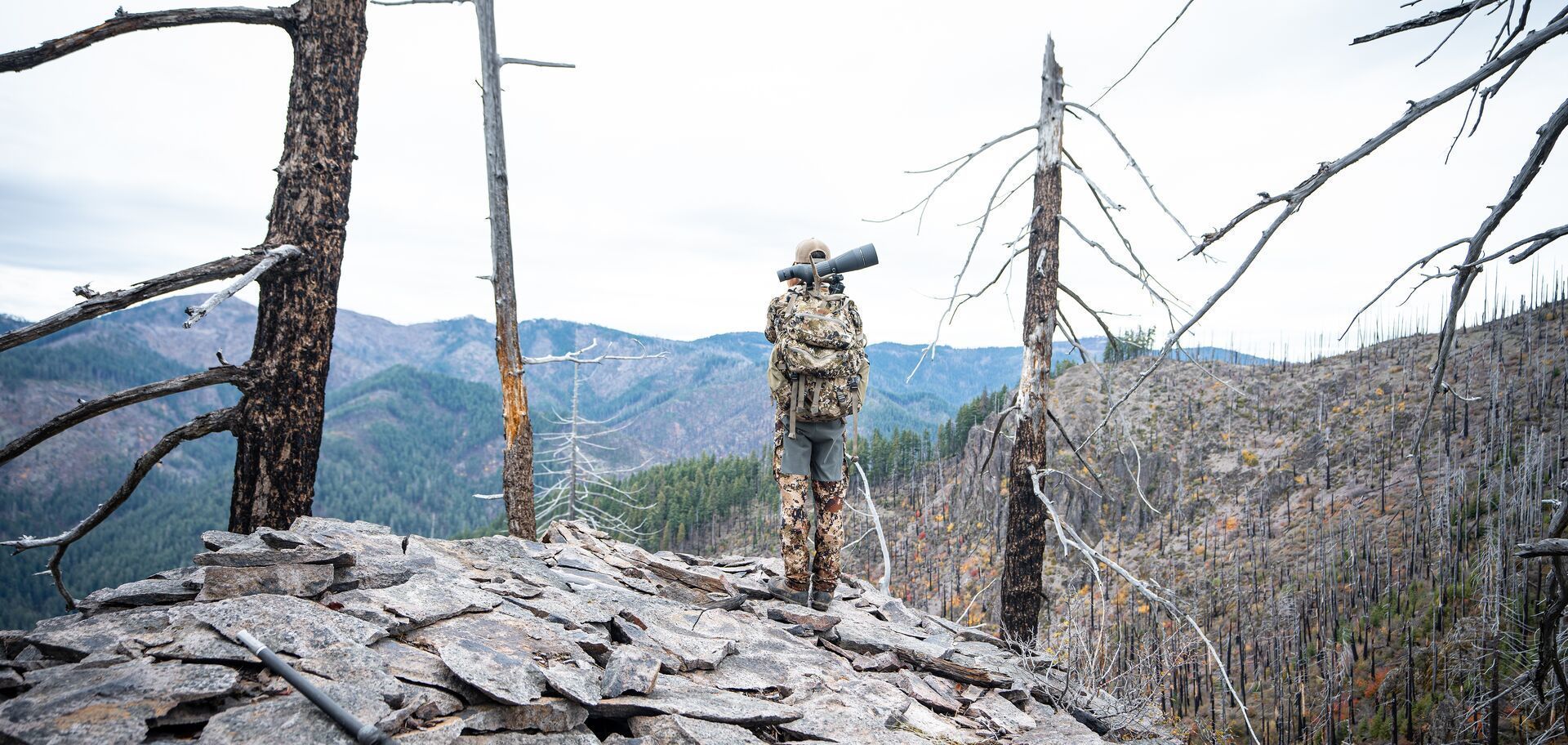 Hunter in camo stands on ridge with hunting gear.