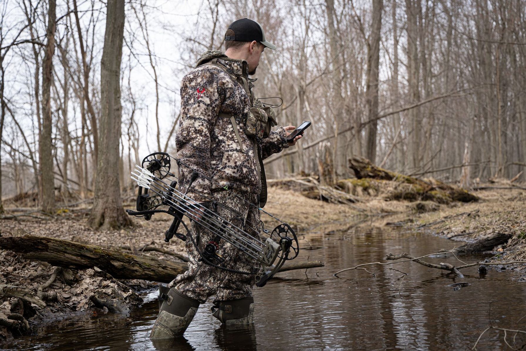 Hunter with bow walks through creek using HuntWise turkey hunting app on phone.