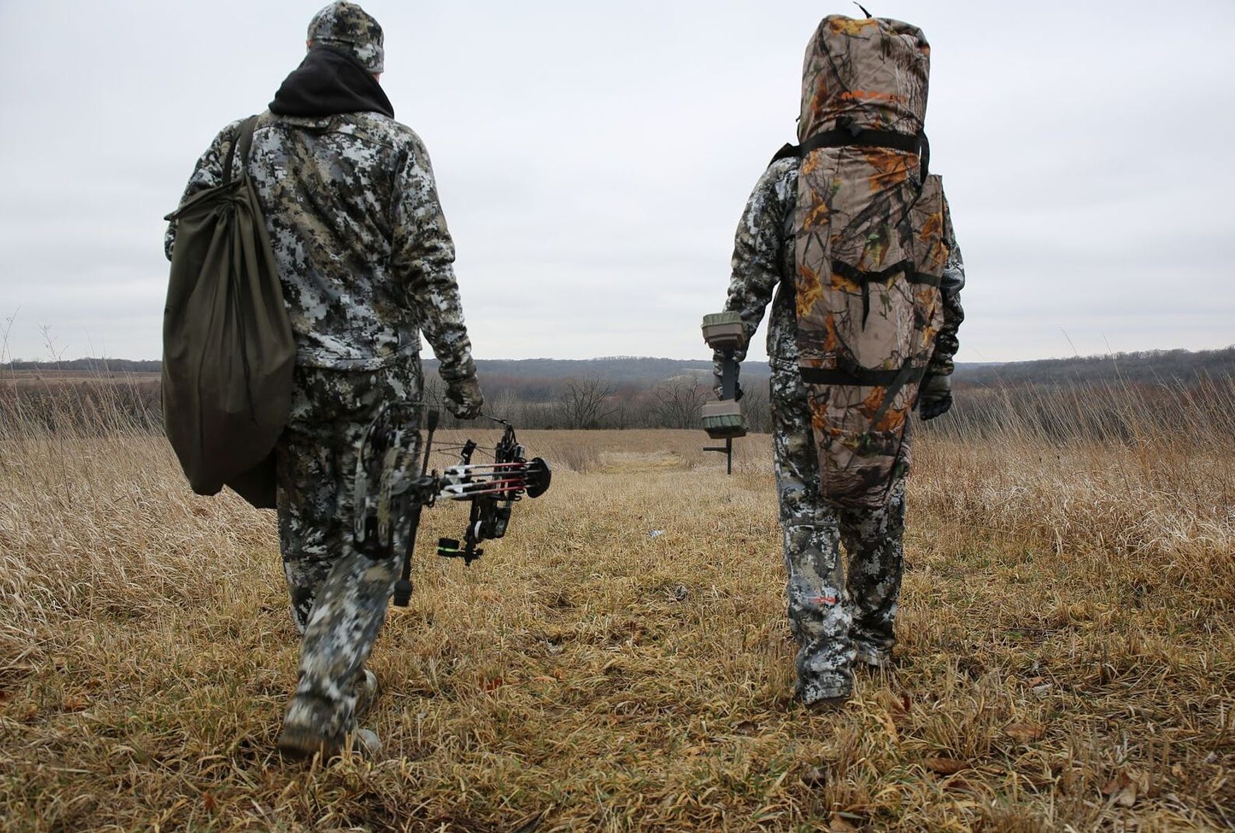 Two hunters wearing hunting packs walking in a field.