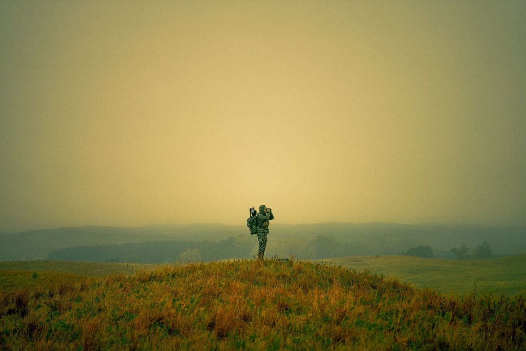 A hunter in the distance on a hill uses binoculars at dusk.
