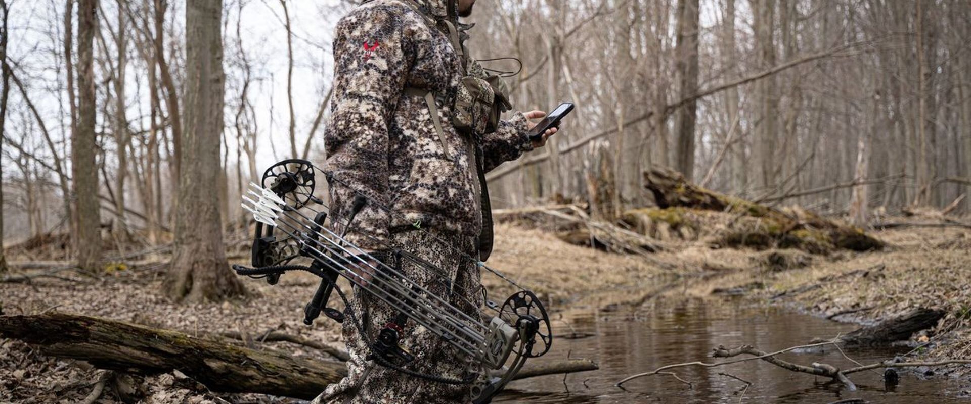 Hunter in the woods with bow holding phone, HuntWise vs Huntstand concept. 