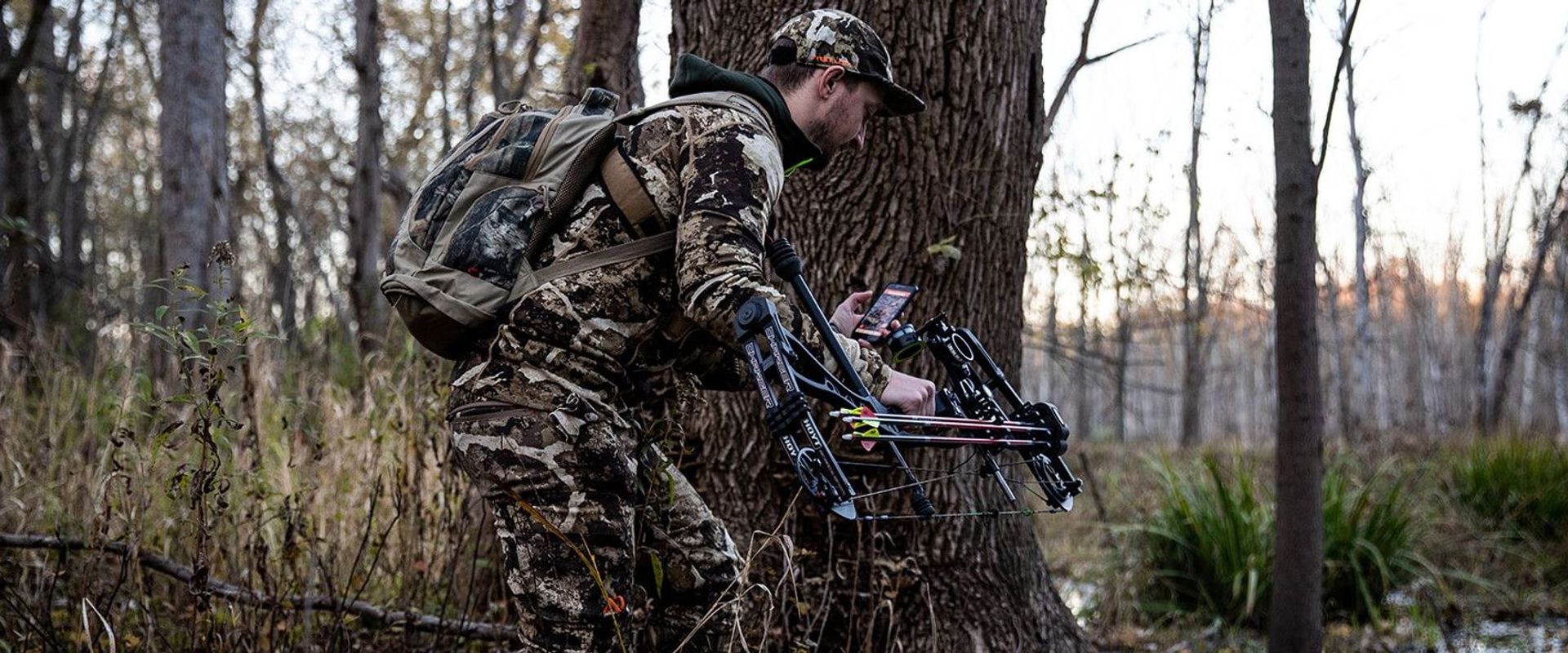 A hunter in camo holding a bow and looking at the HuntWise app on his phone, stalking whitetail concept.