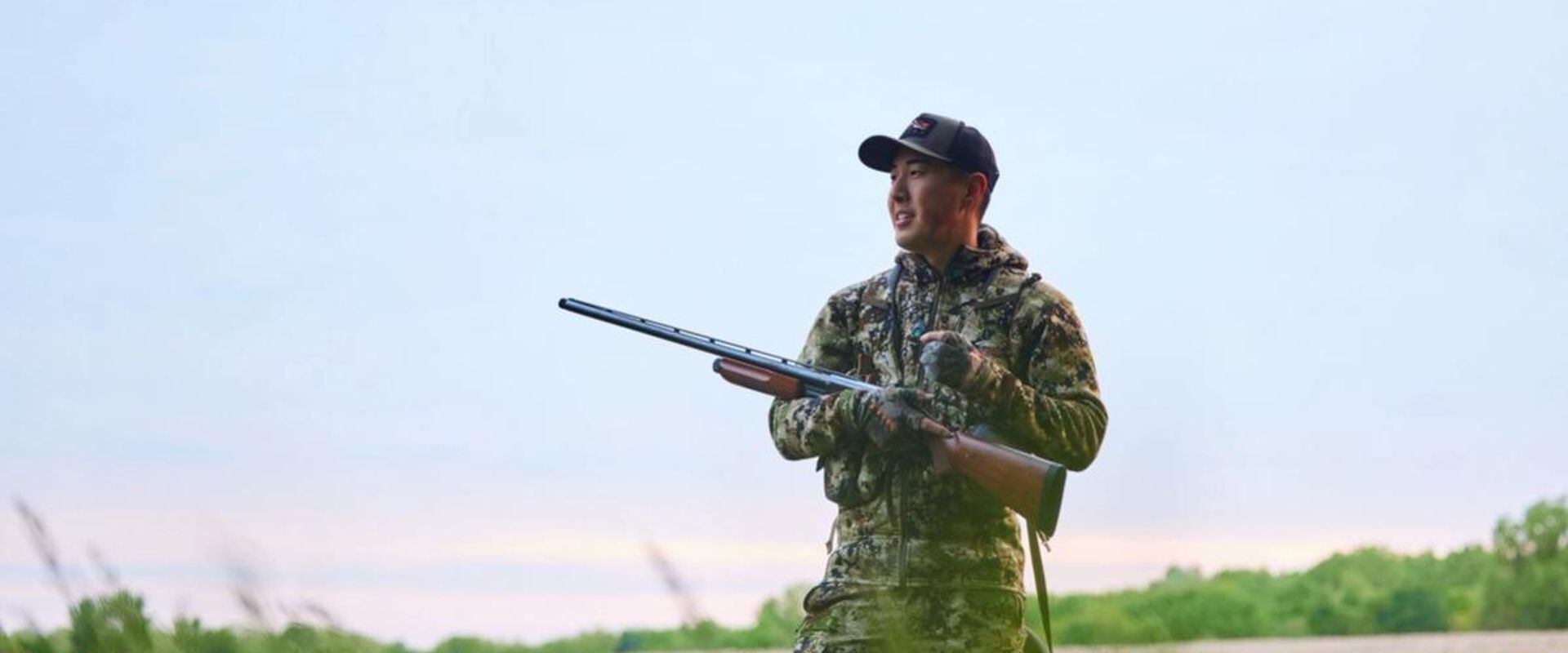 A hunter in camo stands in a field with a firearm, shotgun v rifle concept.