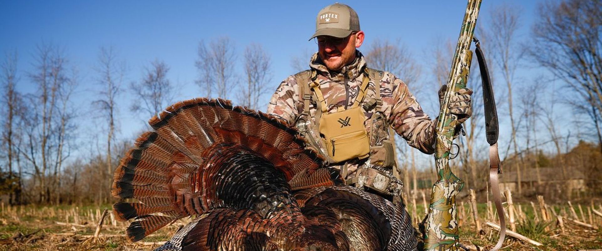 Hunter in camo wearing turkey hunting vest sits with turkey after hunt.