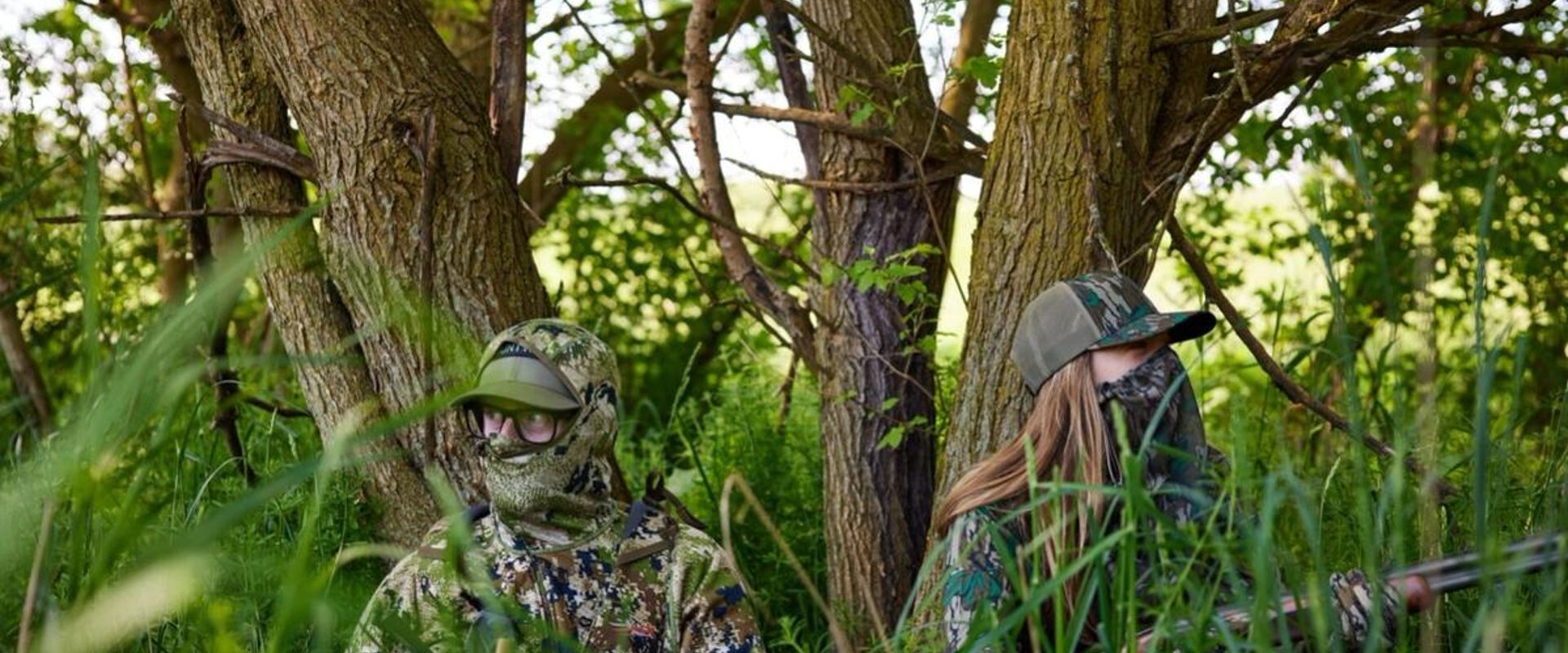 A man and a woman in camo sitting against trees with a shotgun, Osceola turkey hunt concept.