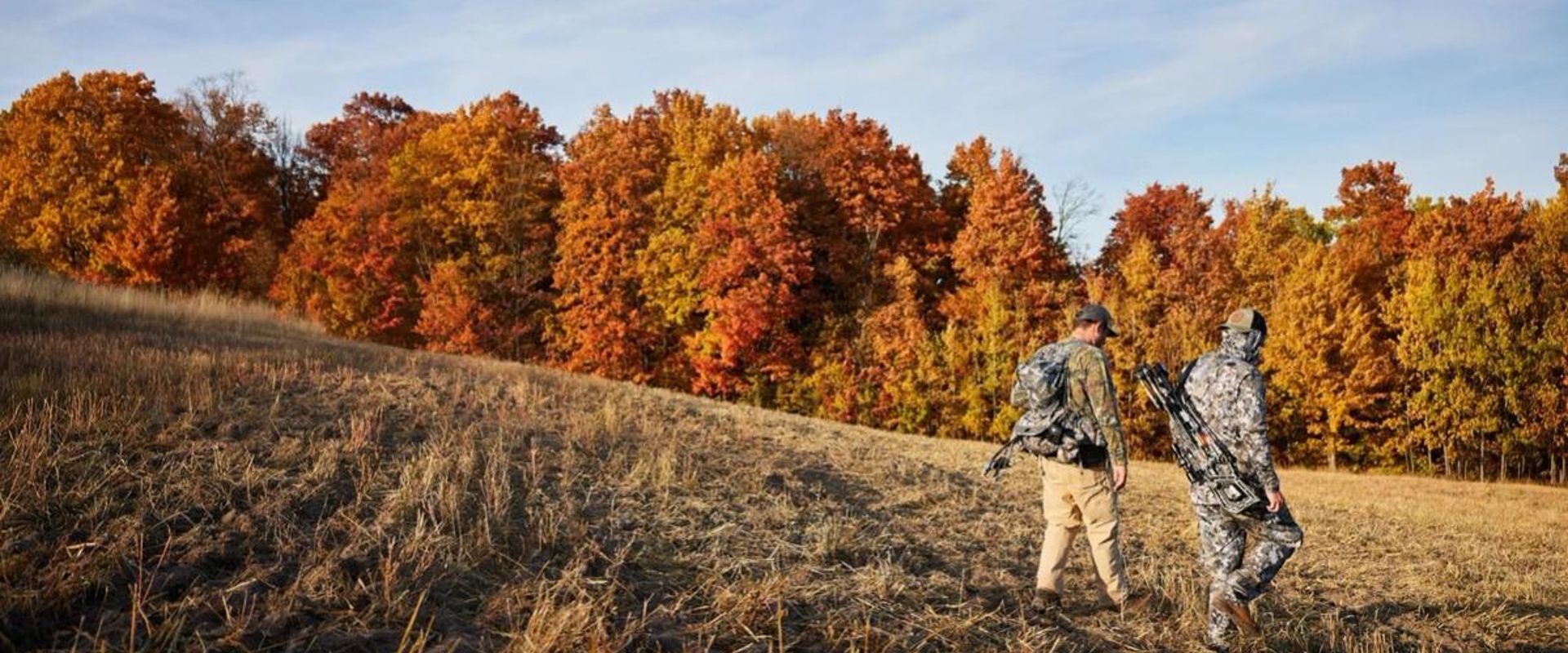 Two hunters walk through a field with hunting gear.