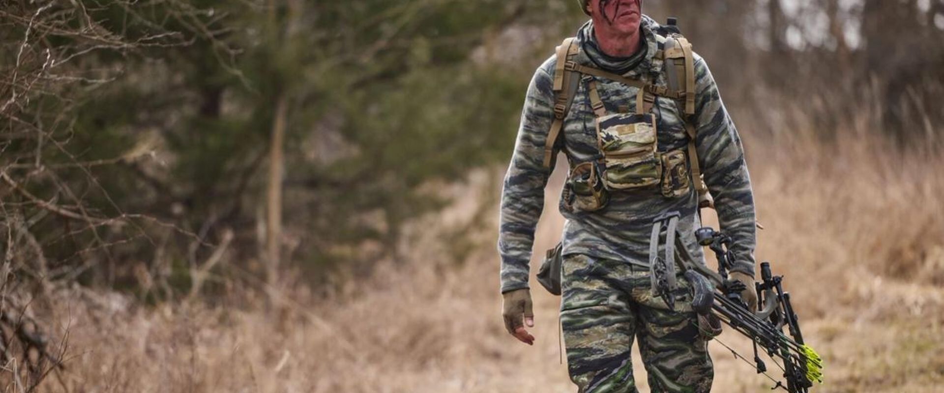John Dudley in hunting camo walks with a bow, bow hunting fitness concept.