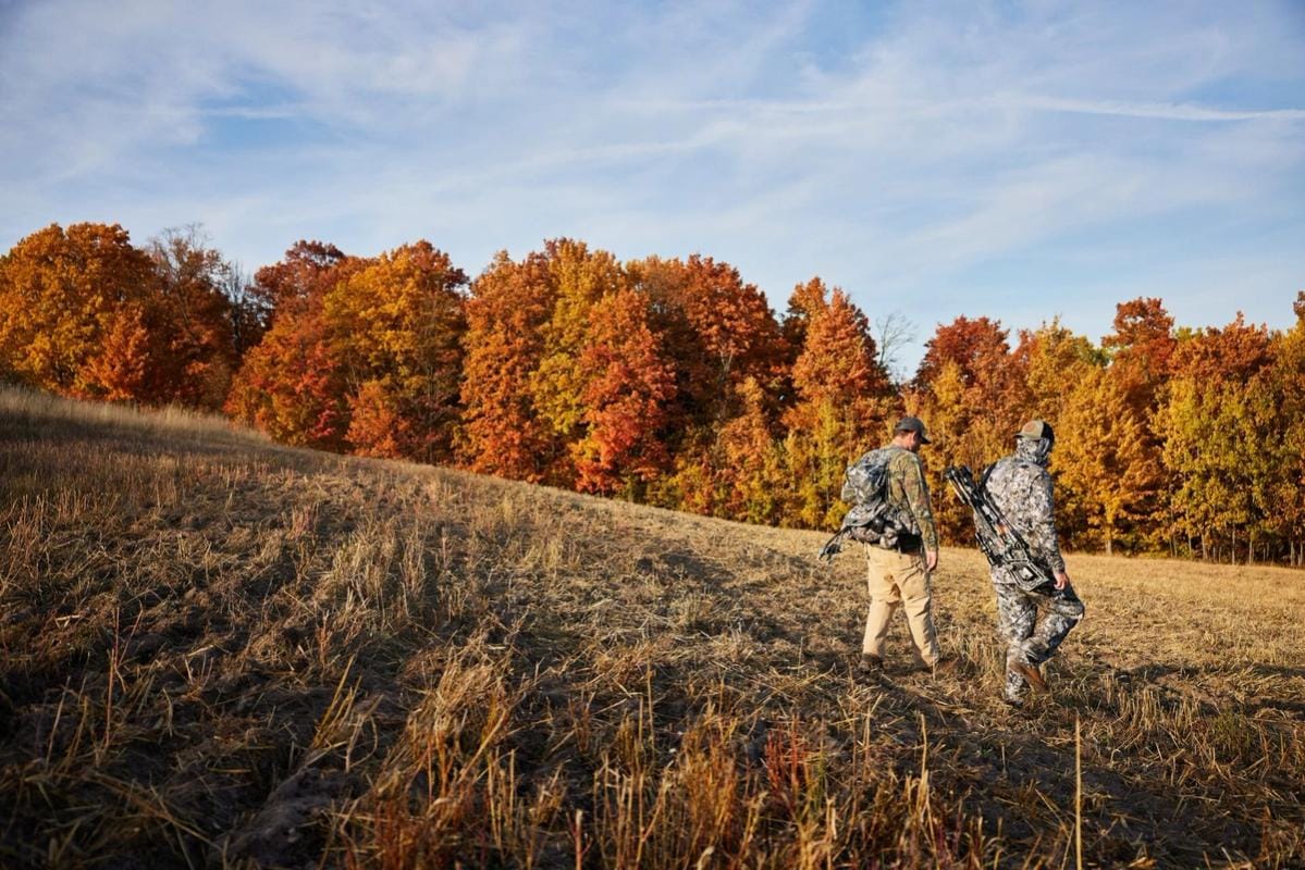 Two hunters walk through a field with hunting gear.