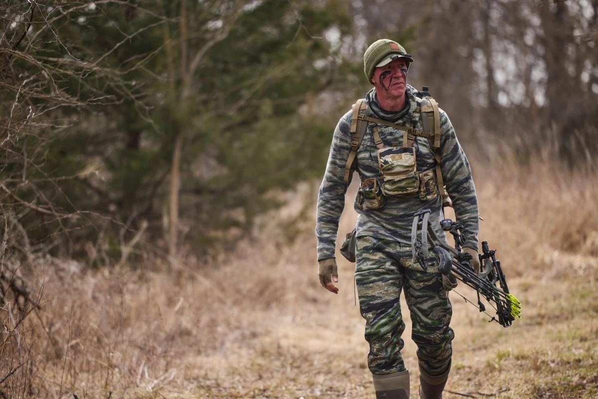 John Dudley in hunting camo walks with a bow, bow hunting fitness concept.