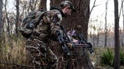 A hunter in camo holding a bow and looking at the HuntWise app on his phone, stalking whitetail concept.
