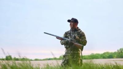 A hunter in camo stands in a field with a firearm, shotgun v rifle concept.