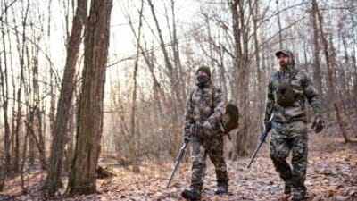Two hunters in turkey gear walk through the woods, hunt Merriam turkey concept.