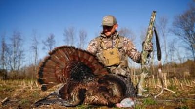 Hunter in camo wearing turkey hunting vest sits with turkey after hunt.