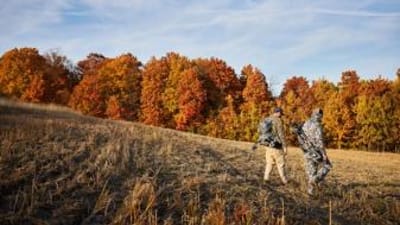 Two hunters walk through a field with hunting gear.