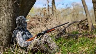 Hunter in camo against tree with shotgun, 12-gauge vs 20-gauge concept.