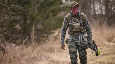 John Dudley in hunting camo walks with a bow, bow hunting fitness concept.