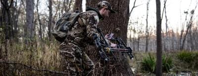 A hunter in camo holding a bow and looking at the HuntWise app on his phone, stalking whitetail concept.