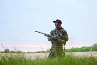 A hunter in camo stands in a field with a firearm, shotgun v rifle concept.