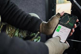 Close-up of hands holding a phone showing the HuntWise hunting weather app on screen.
