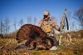 Hunter in camo wearing turkey hunting vest sits with turkey after hunt.