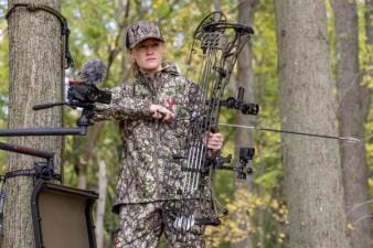 A female hunter in a tree stand gets ready to aim a bow, gun vs. bow shot placement concept.