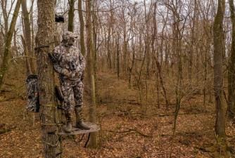 A hunter in camo in a tree stand looks out into the woods.