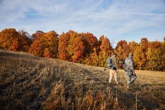 Two hunters walk through a field with hunting gear.