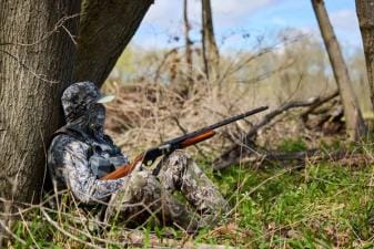 Hunter in camo against tree with shotgun, 12-gauge vs 20-gauge concept.