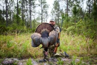 Hunter kneels with turkey after hunt, best caliber for turkey hunting concept.