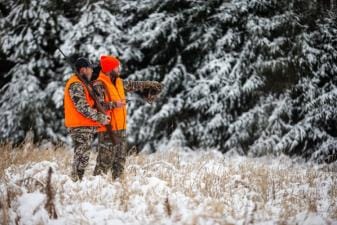 Two hunters in blaze orange look into snowy trees, late-season whitetail hunting concept.