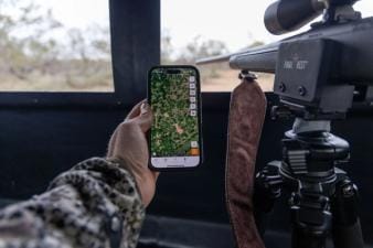 Hunter holds phone showing Huntwise next to rifle in blind, where can I hunt concept.